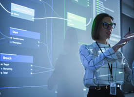 A woman confidently delivering a presentation in front of a projector screen displaying visuals. The room is dark with blue lighting, highlighting her presence as she engages the audience with gestures, showcasing her expertise and poise.