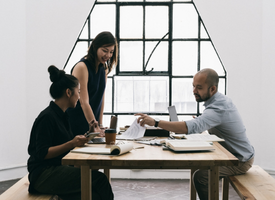 Coworkers seated around a bench and table in a bright, white office, actively discussing a project. The scene is clean and modern, with natural light enhancing the workspace.