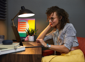A woman working on a laptop in a cozy home office, seated at a desk with a stylish lamp. She is reclining her head on one hand, appearing focused and relaxed.